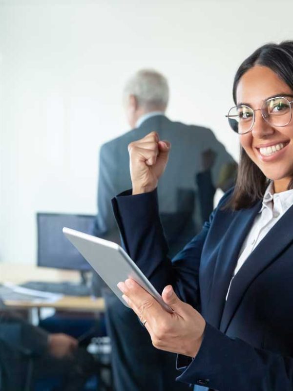 feliz-mujer-profesional-gafas-traje-sosteniendo-tableta-haciendo-gesto-ganador-mientras-dos-hombres-negocios-trabajan-detras-pared-vidrio-copie-espacio-concepto-comunicacion